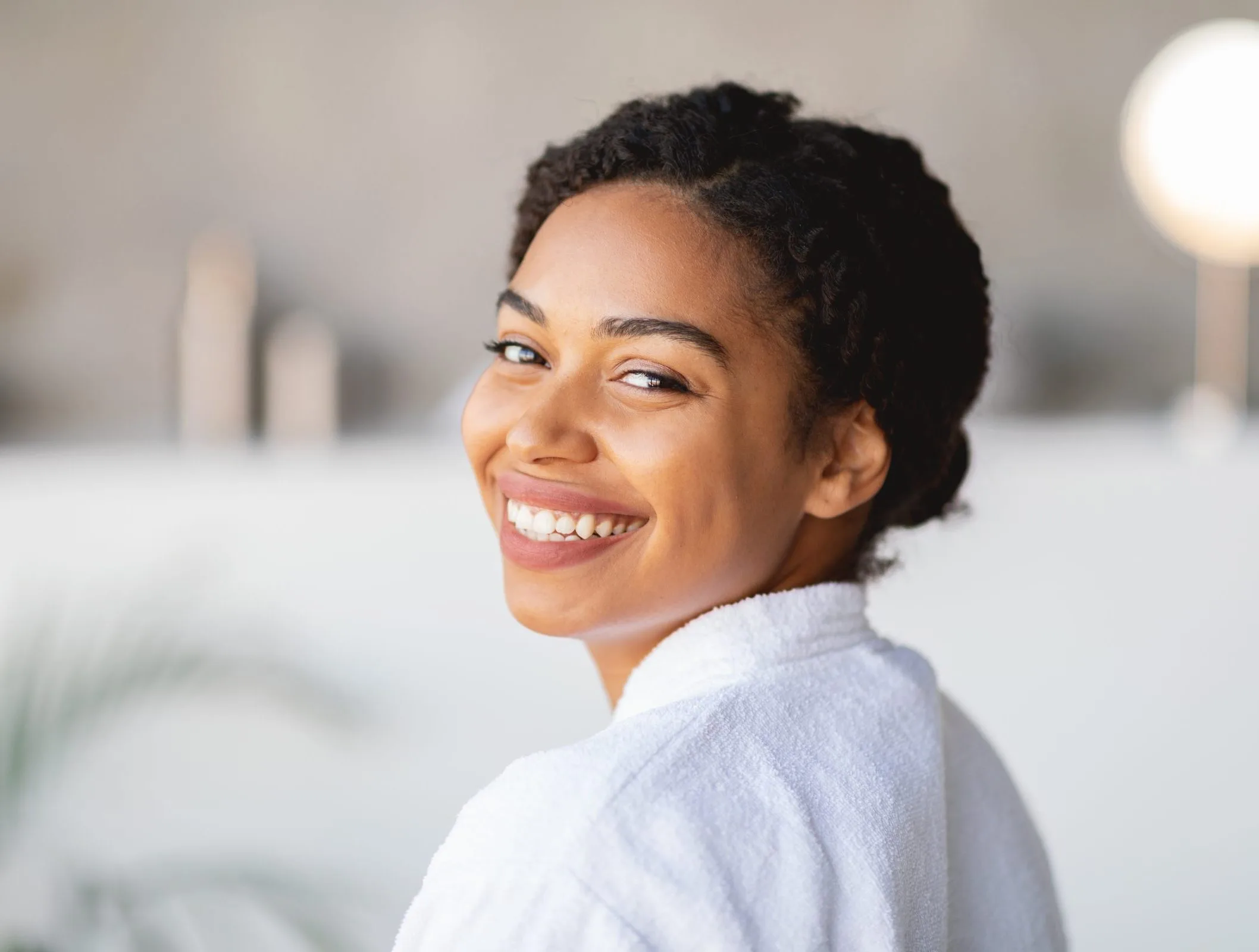 A smiling woman with curly hair wearing a white robe in a bright, modern spa-like interior