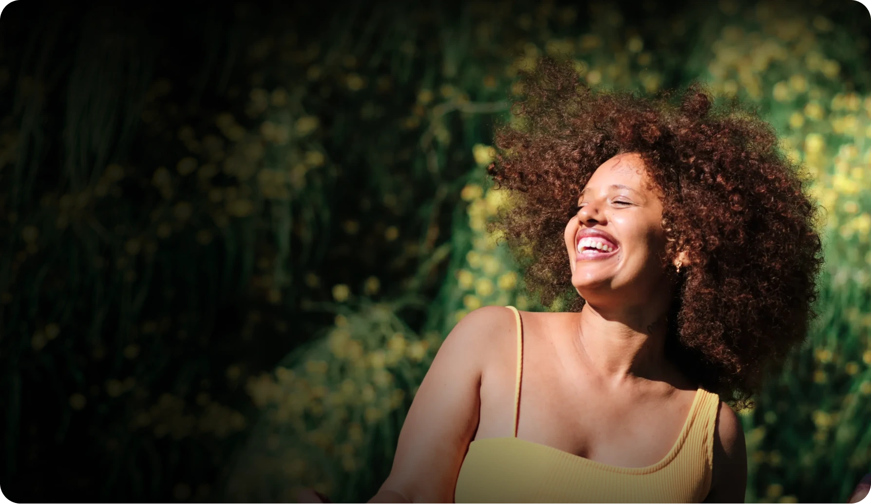 A joyful woman with curly hair laughs with her eyes closed against a sunlit natural backdrop of green foliage