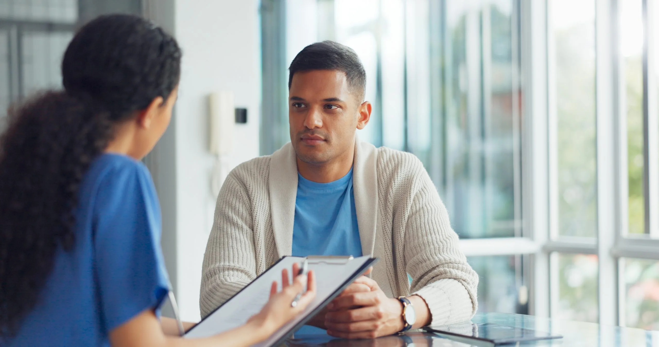 A male patient listens attentively during a consultation with a provider holding a clipboard in a bright offic