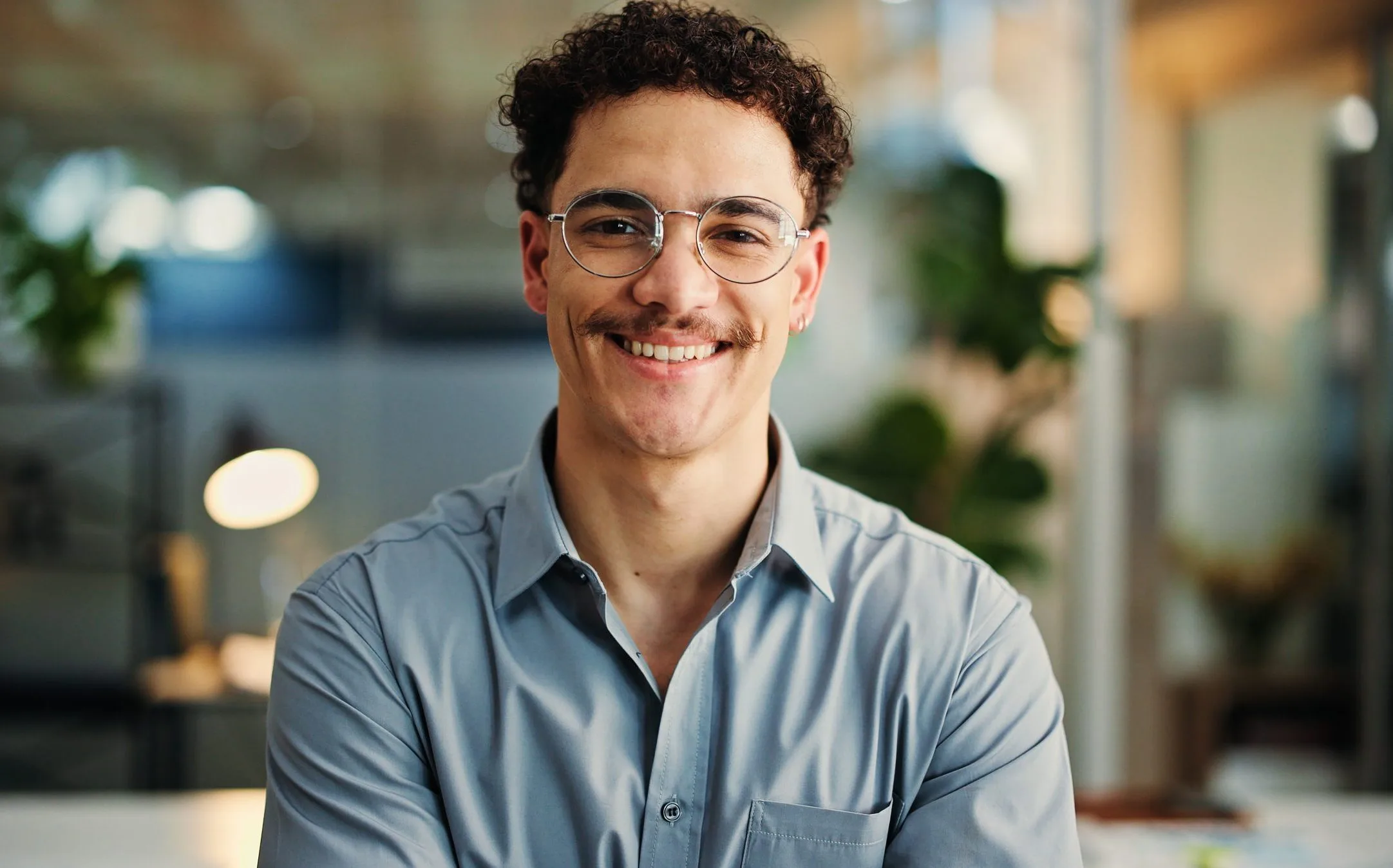 A smiling young man with curly hair and glasses in a modern office setting