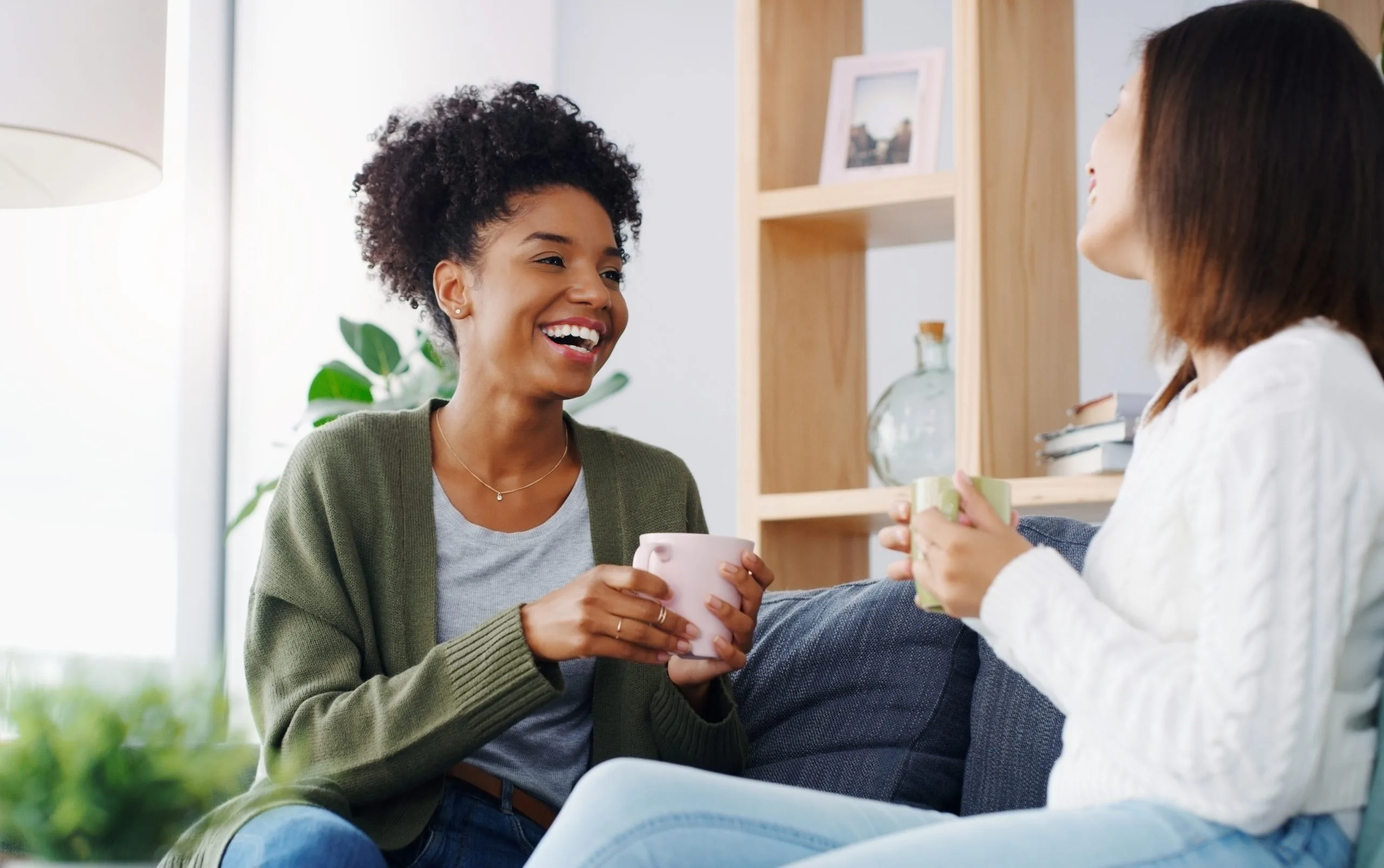 Two women laughing and conversing while holding coffee cups in a bright, modern living room