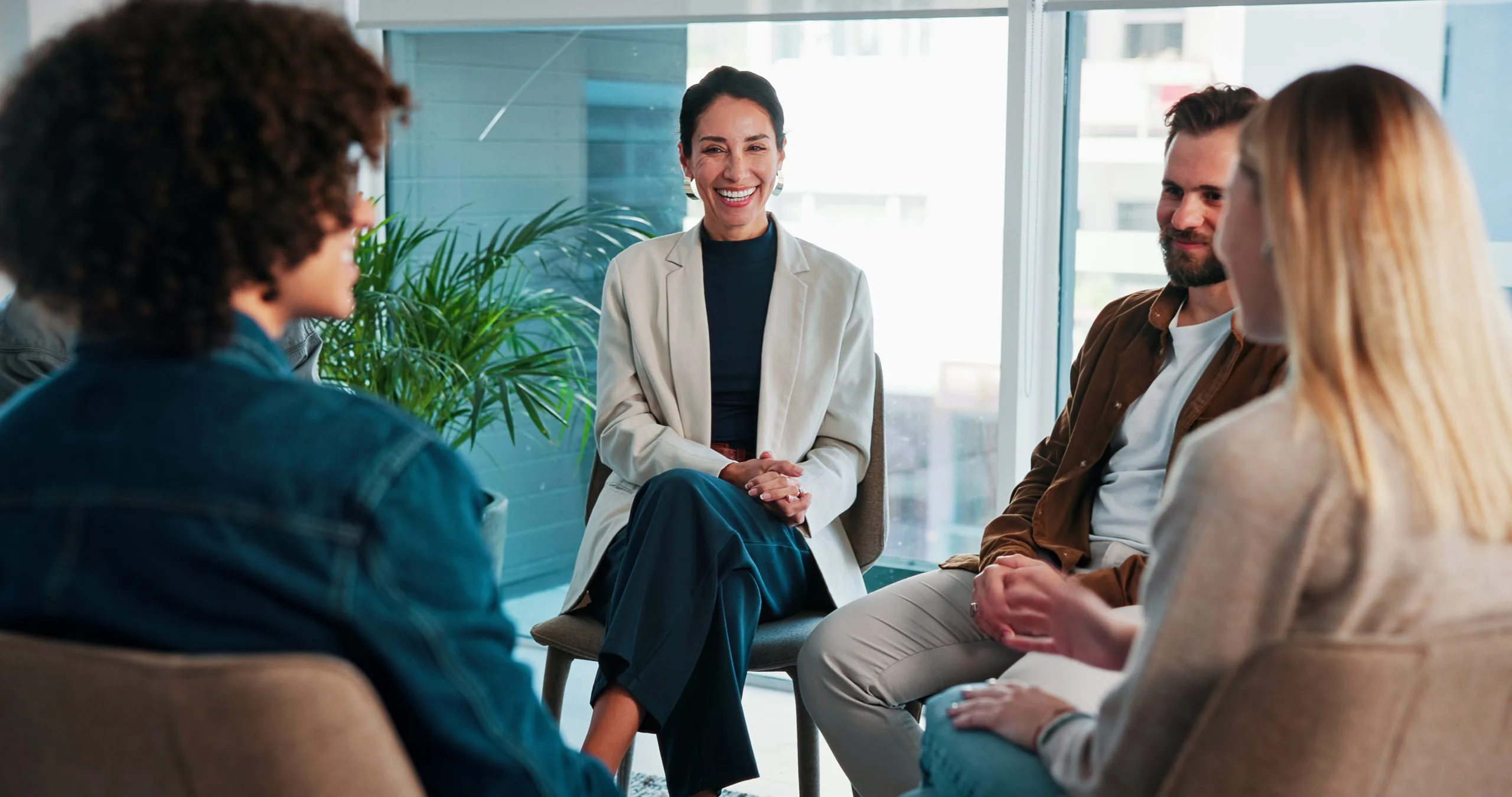 Group therapy session with five people sitting in a circle in a bright office with windows and plants