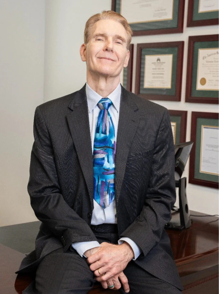 Man in dark suit and blue tie sitting at desk with framed credentials on wall behind him