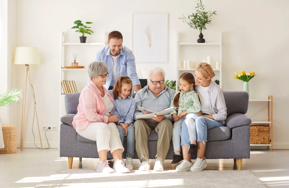 Multi-generational family sitting on a gray sofa, enjoying a storybook together in a bright living room