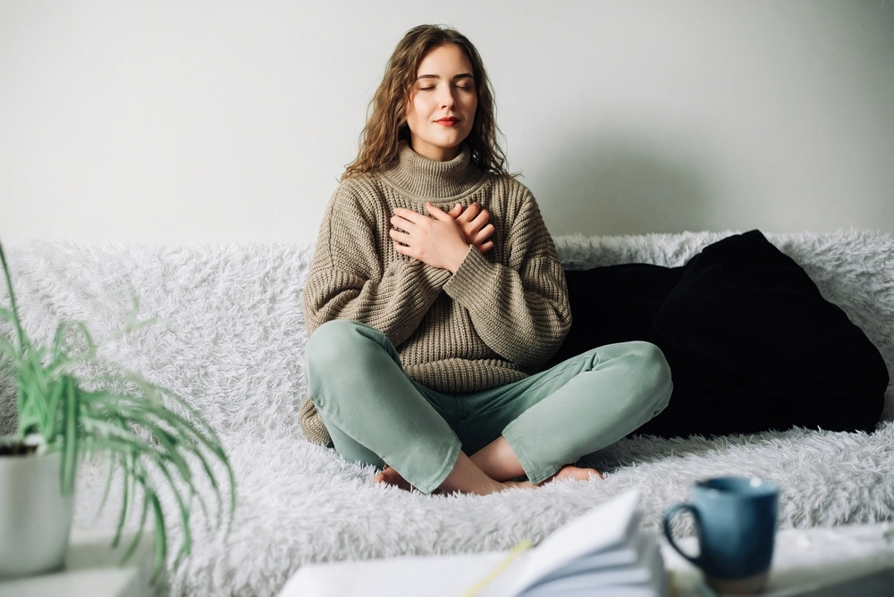 Woman meditating on a white fuzzy sofa with hands over her chest, eyes closed, in a calm living room