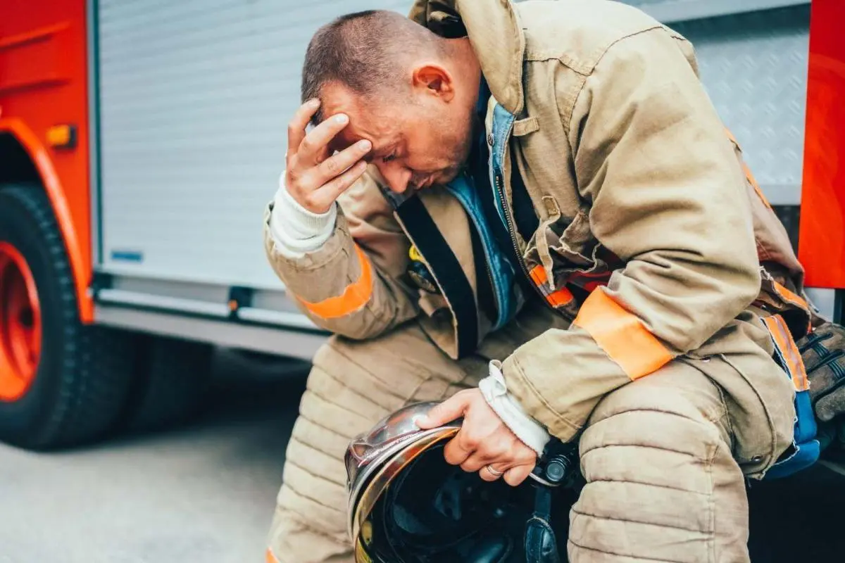 Firefighter sitting on fire truck steps holding helmet, looking stressed