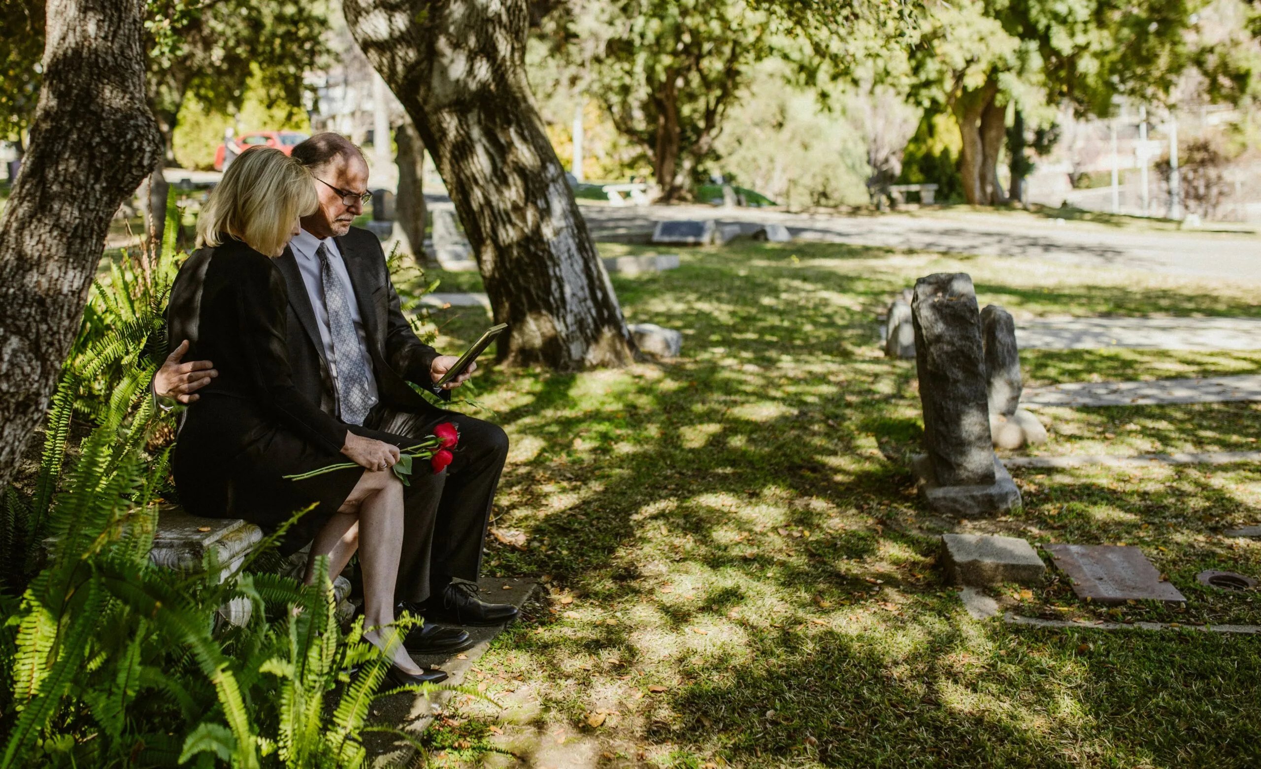 Mourning couple sitting at cemetery looking at photo while holding roses