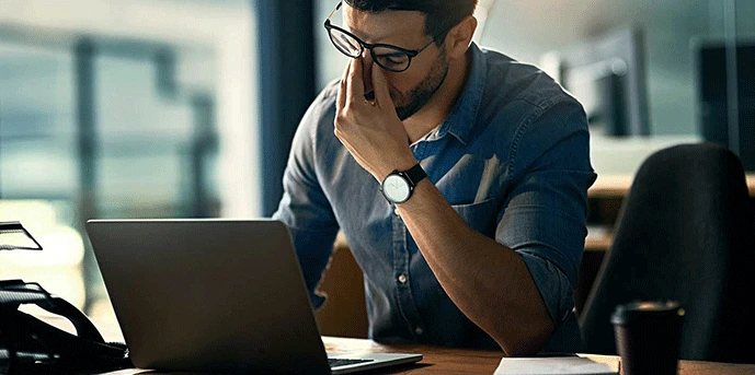 Man in office sitting at laptop, pinching nose in stress or fatigue