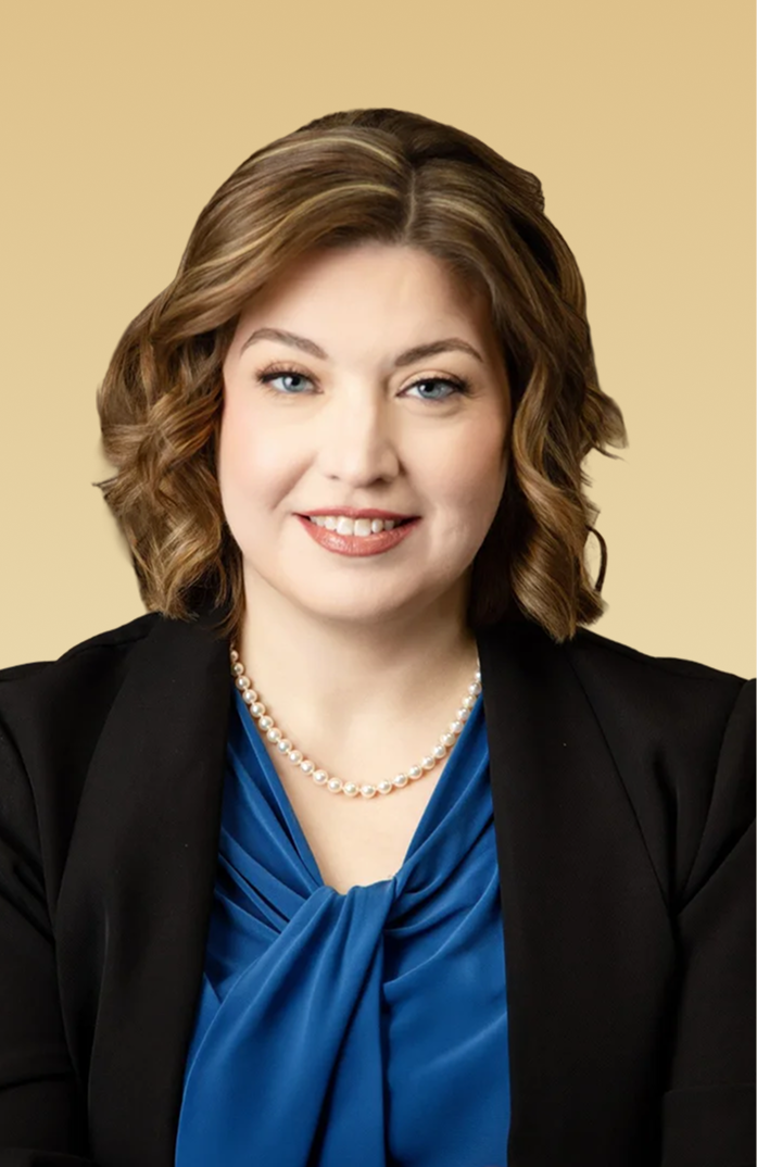 Professional headshot of a woman with brown wavy hair wearing a black blazer and blue top against a tan background