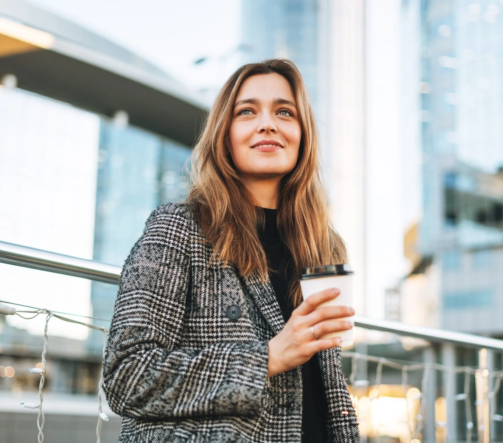 Woman in plaid blazer looking upward on modern building balcony with city backdrop