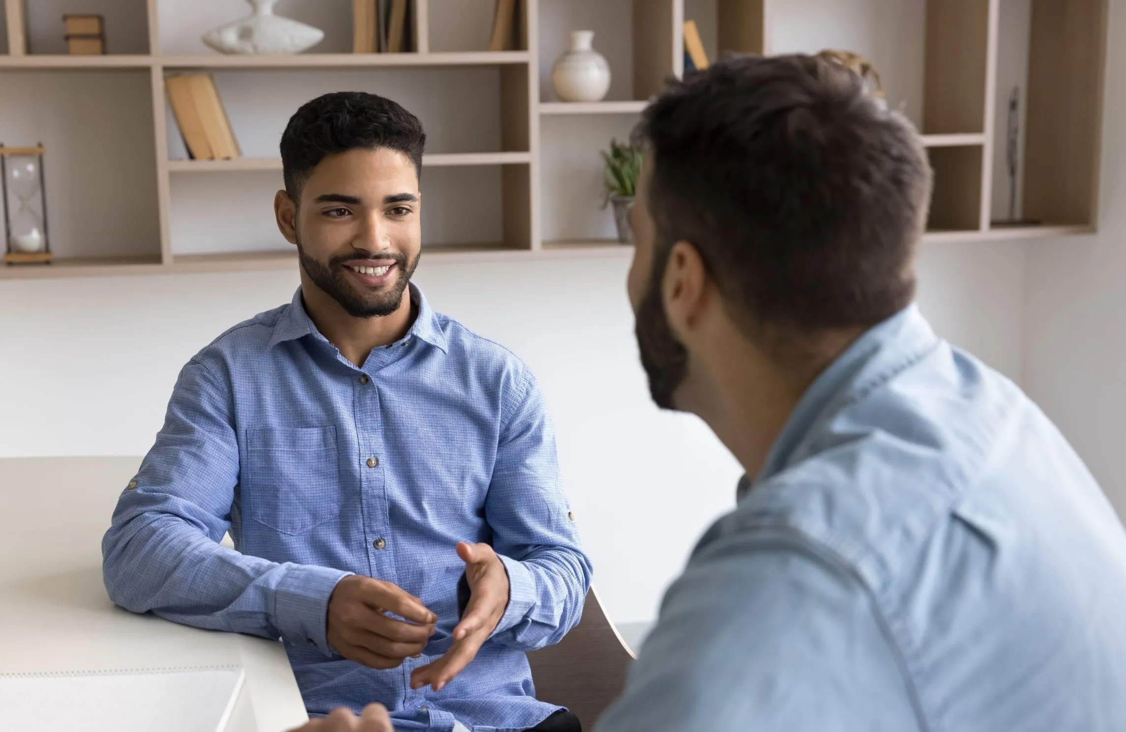 Male therapist in blue shirt speaking with a patient across a white desk in a modern office
