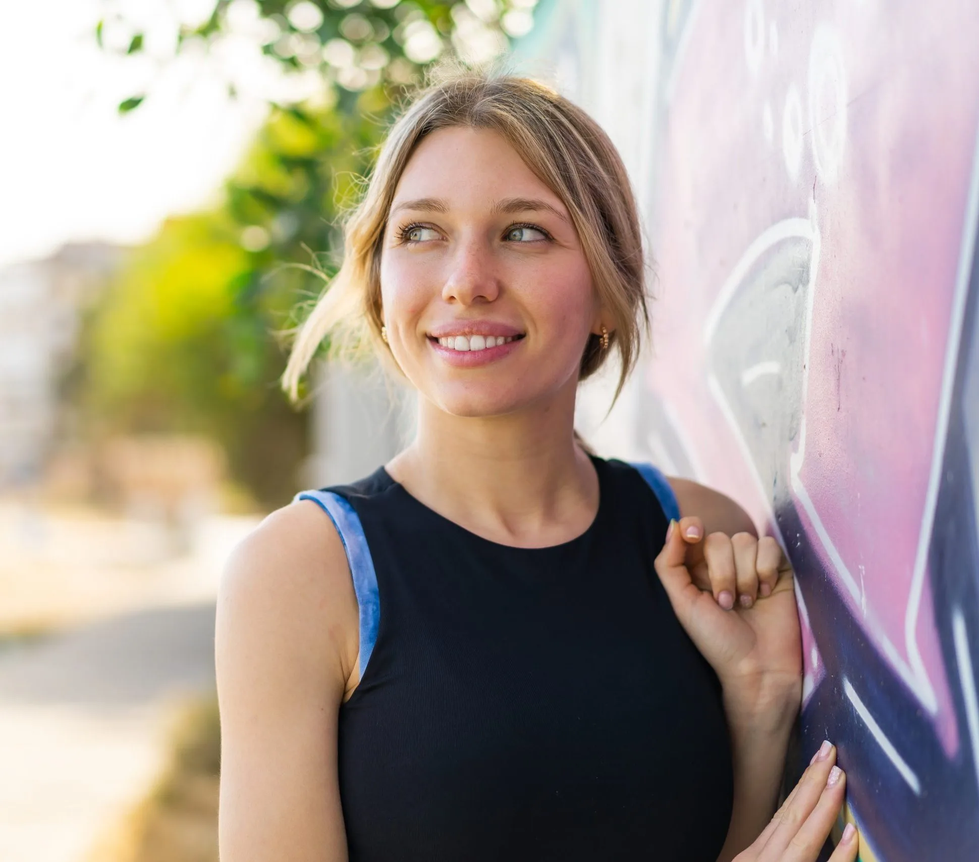 Smiling woman standing outdoors against a pink wall with green foliage in background