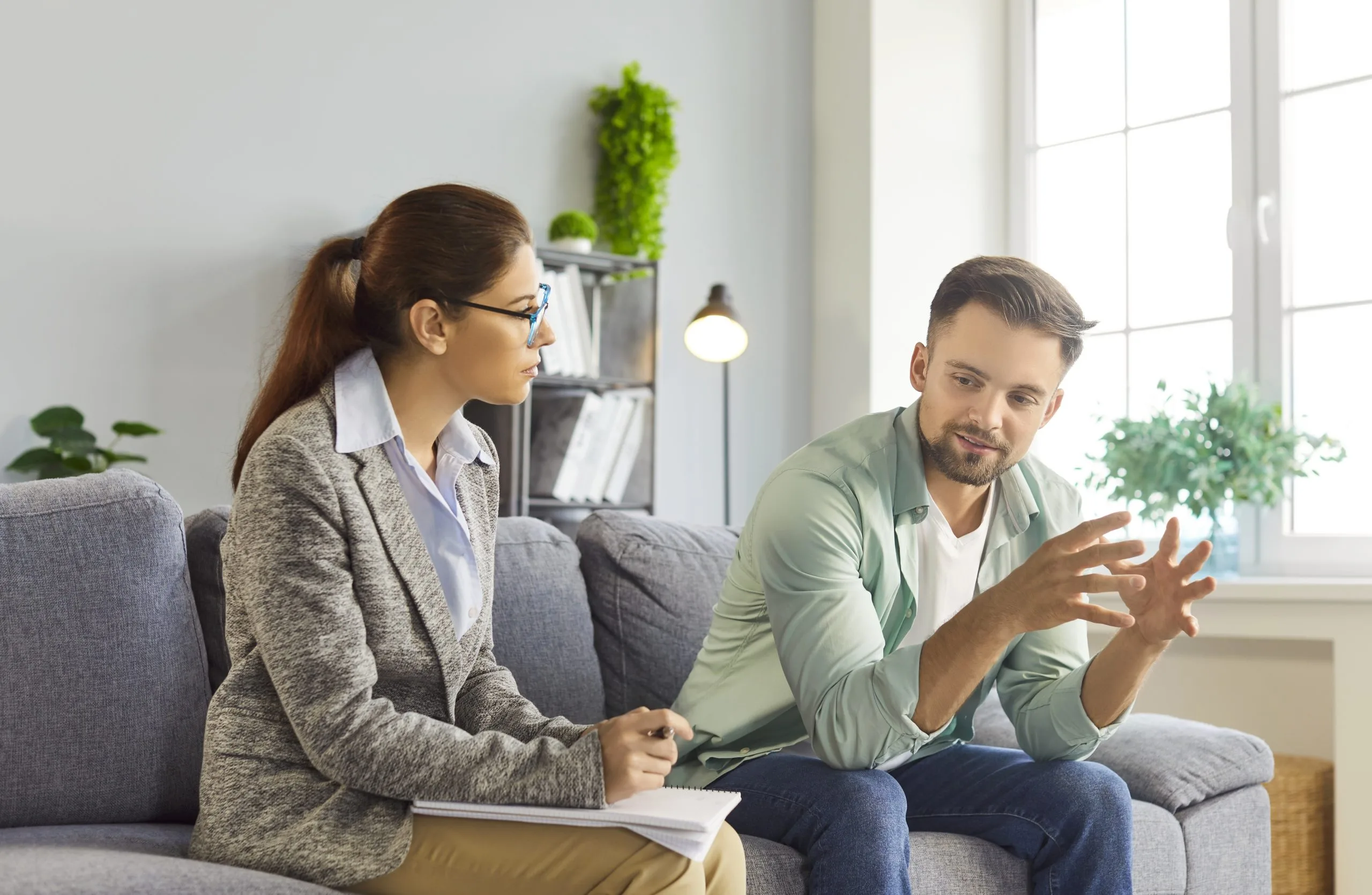 Therapist taking notes while listening to a male patient speaking during a counseling session in a bright, plant-filled office