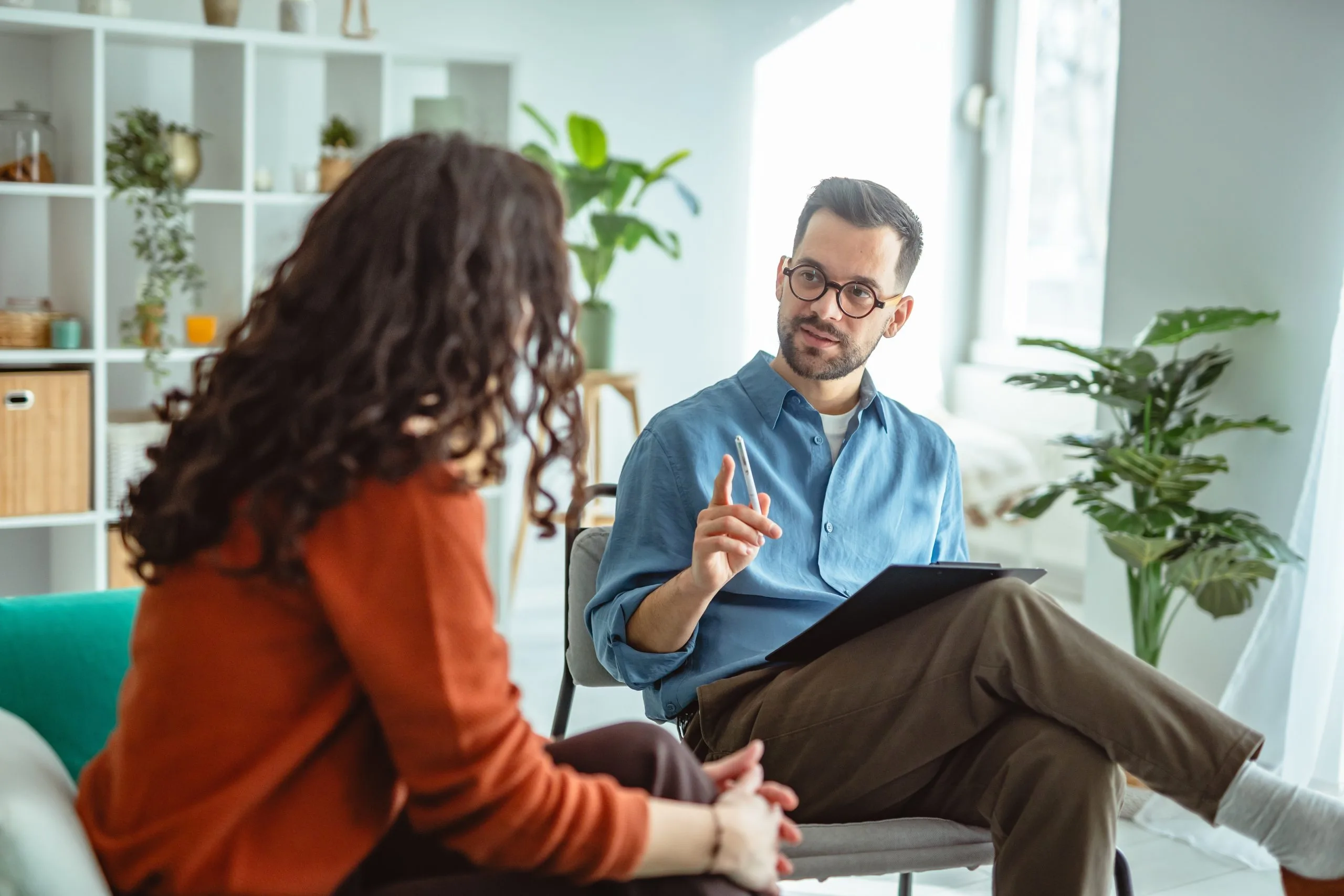 Therapist in blue shirt discussing notes with patient in bright office with plants