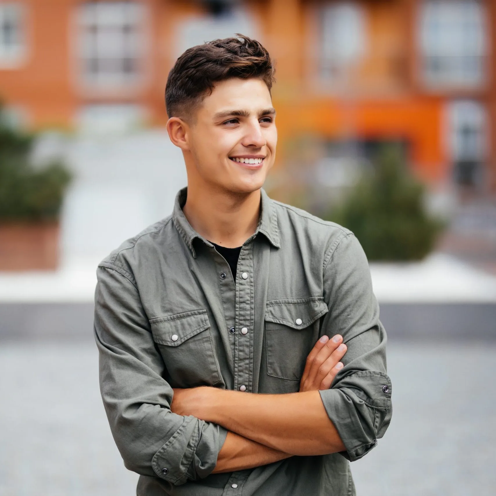 Young man smiling with arms crossed outdoors in front of a brick building