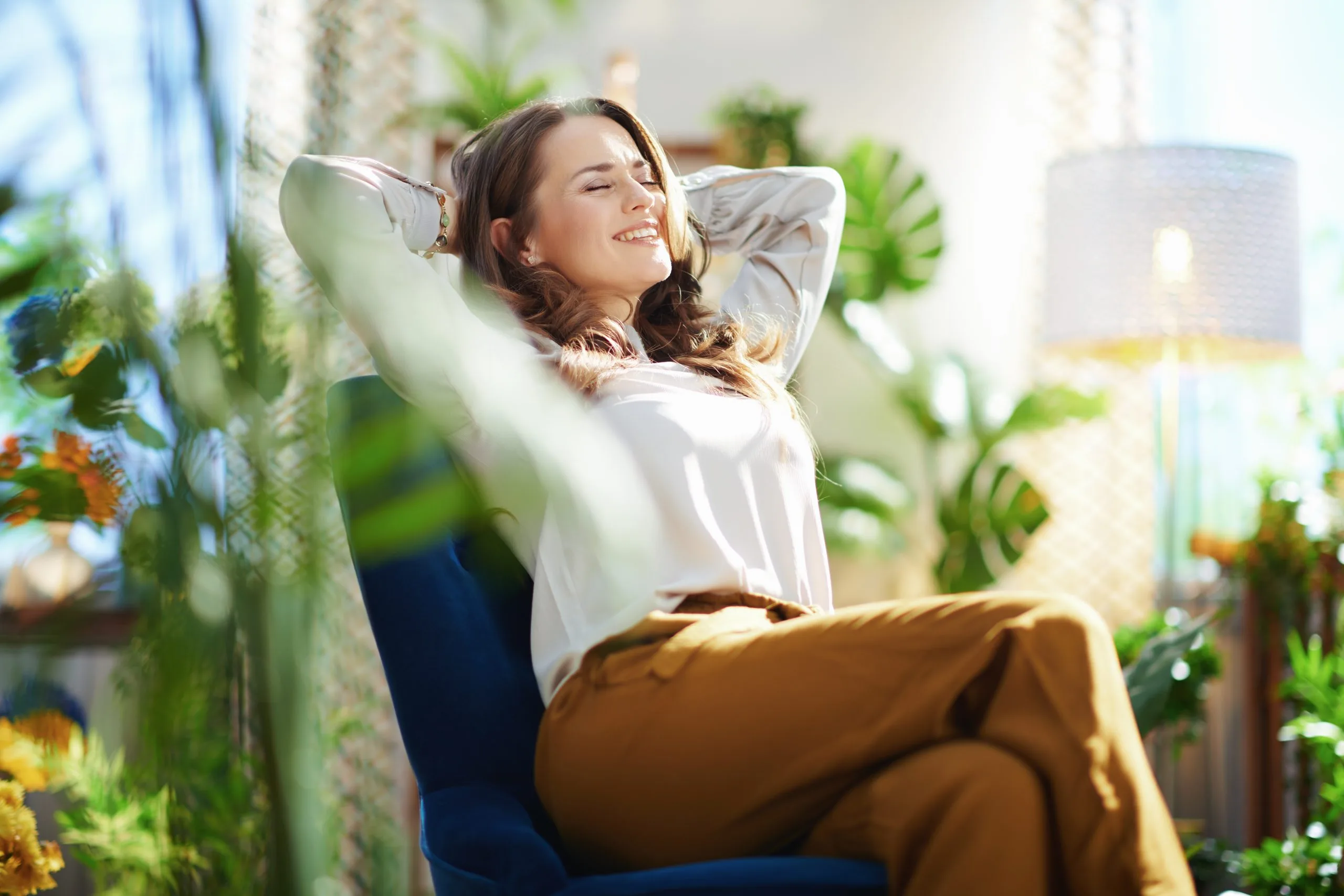 Woman in white shirt relaxing in chair surrounded by green plants with arms behind her head