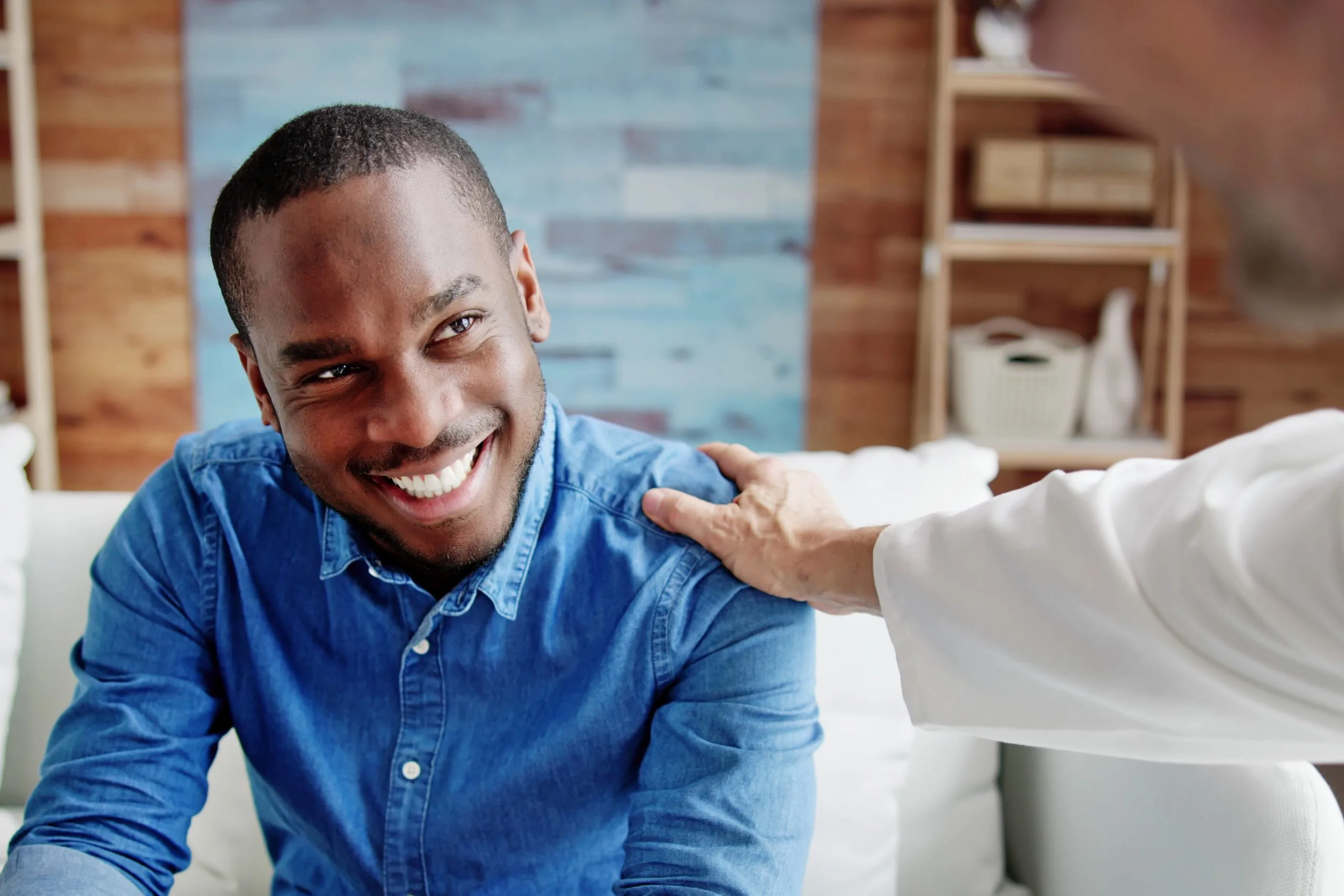 Smiling man in denim shirt receiving supportive hand on shoulder from healthcare provider in clinical setting