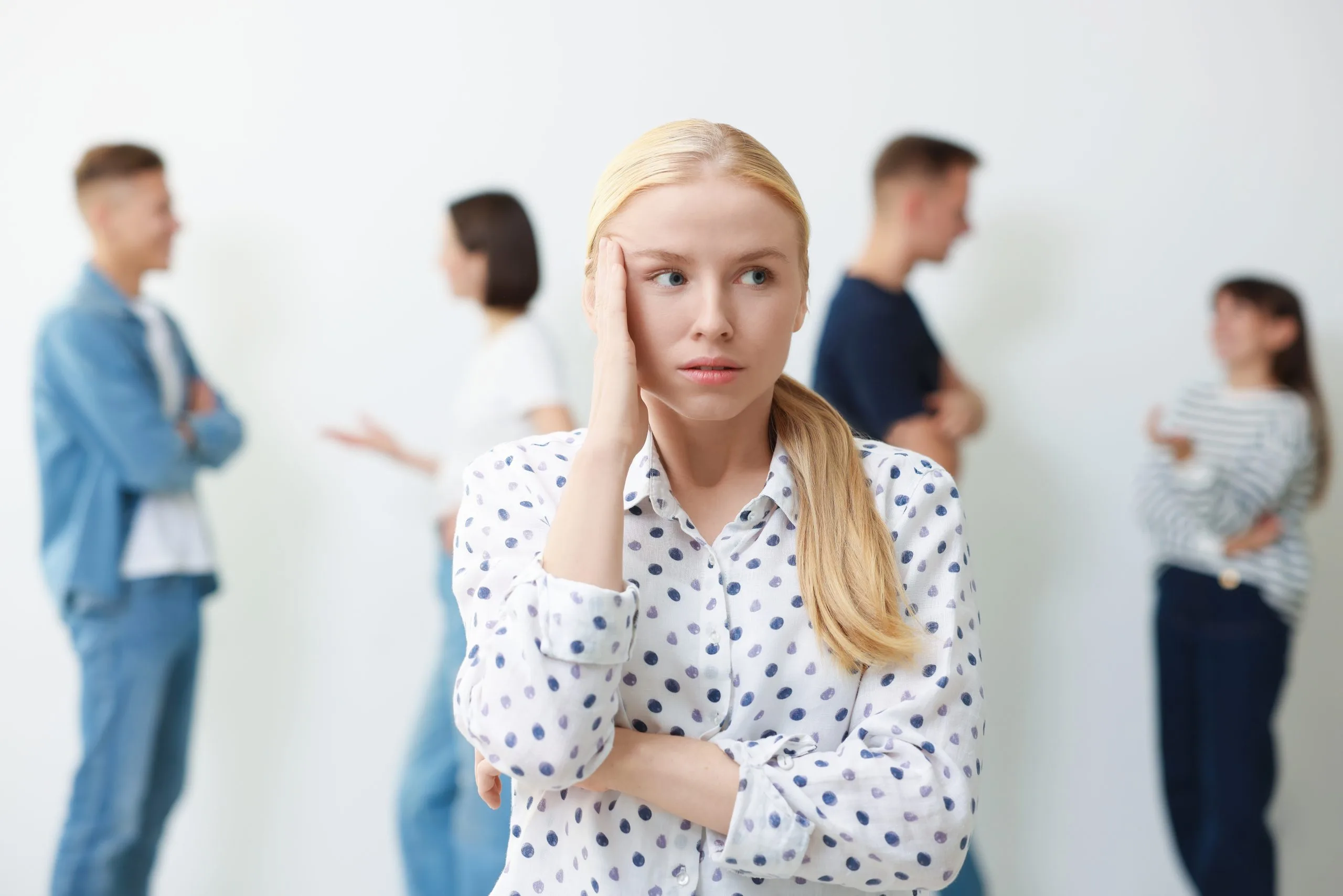 Blonde woman in polka-dot shirt looking worried with hand to face, other people conversing blurred in background