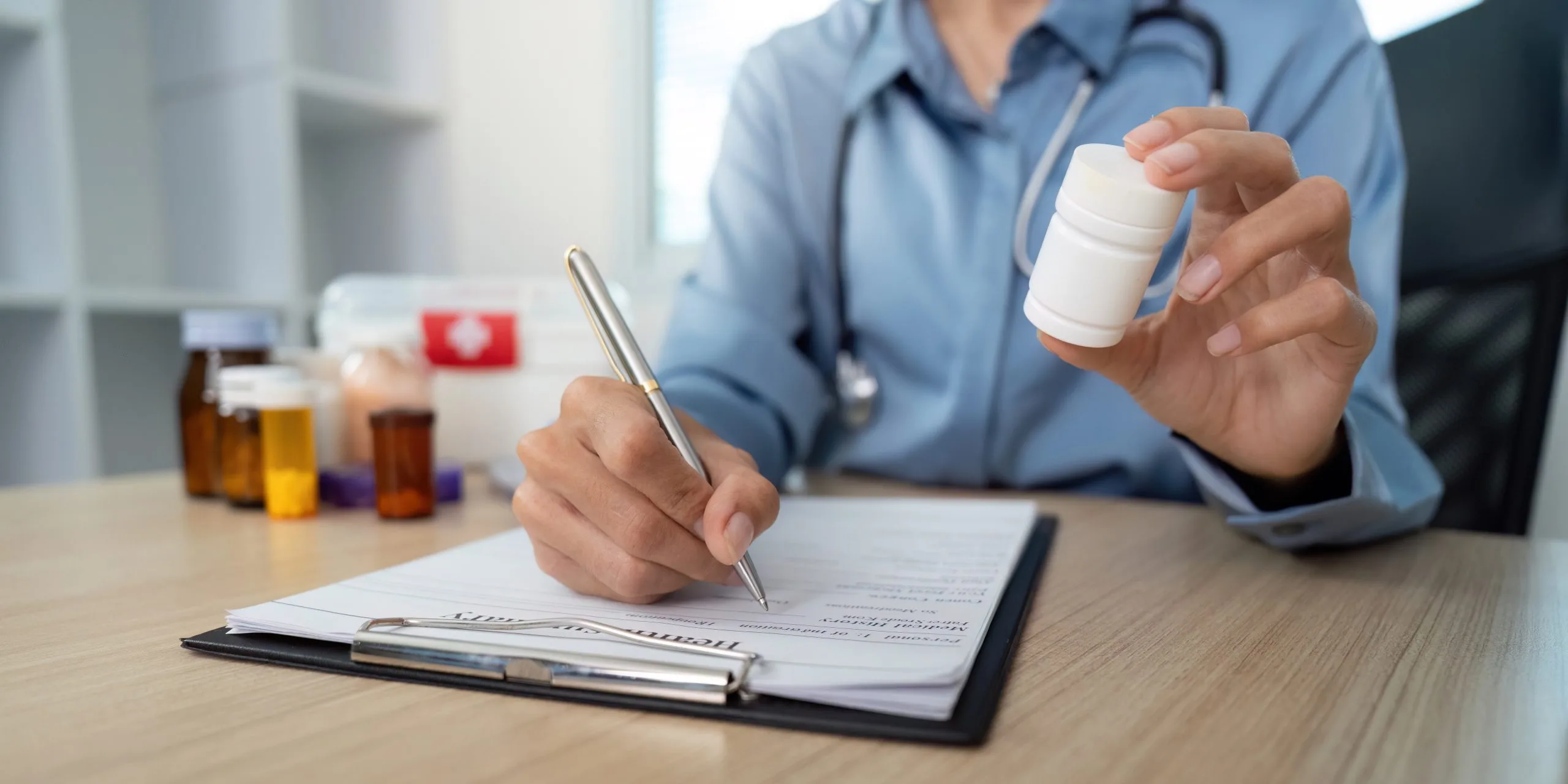 Healthcare provider in blue scrubs holding medication bottle while writing in patient chart at desk with medicine bottles nearby