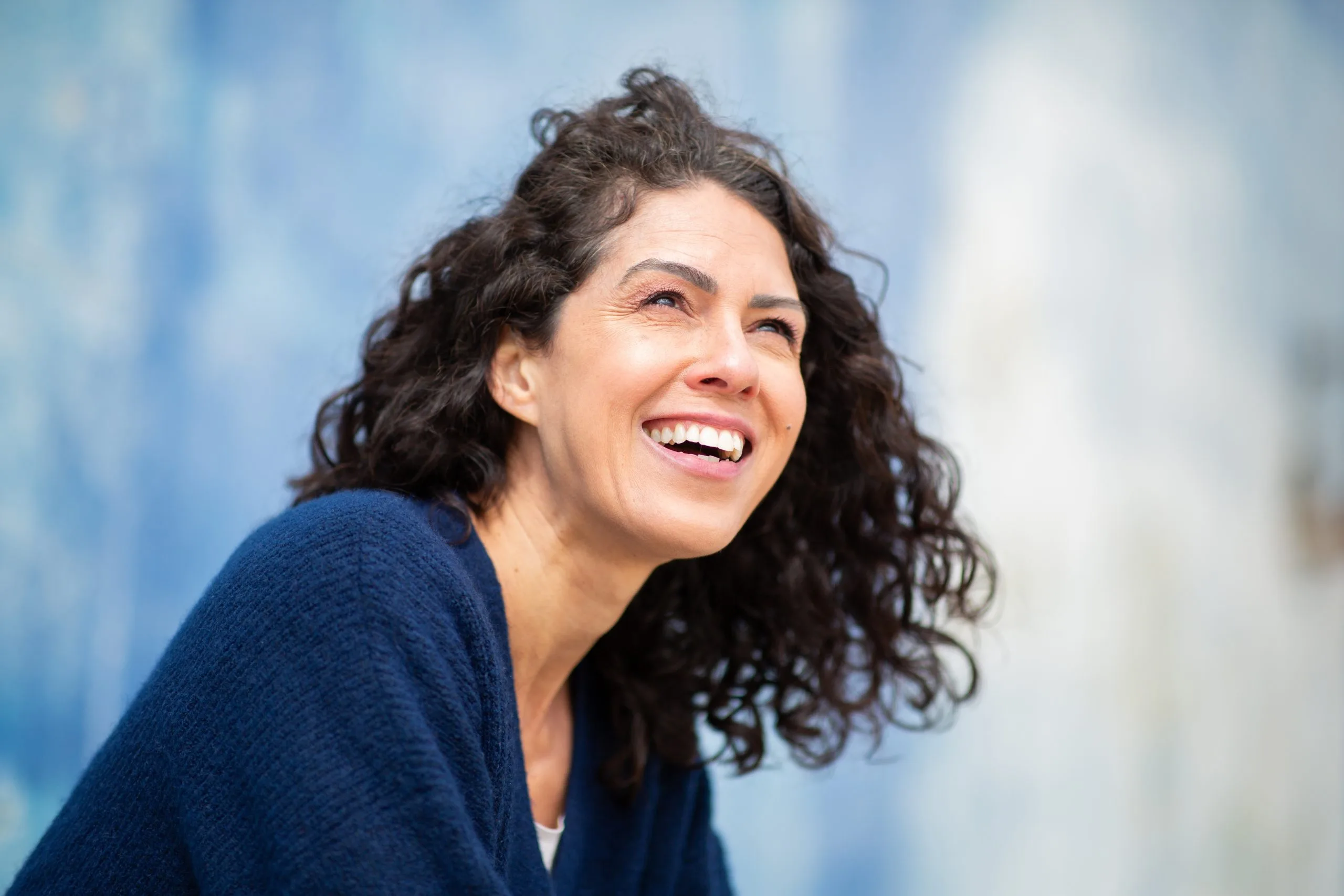 Woman with curly dark hair in navy sweater laughing against blue textured wall