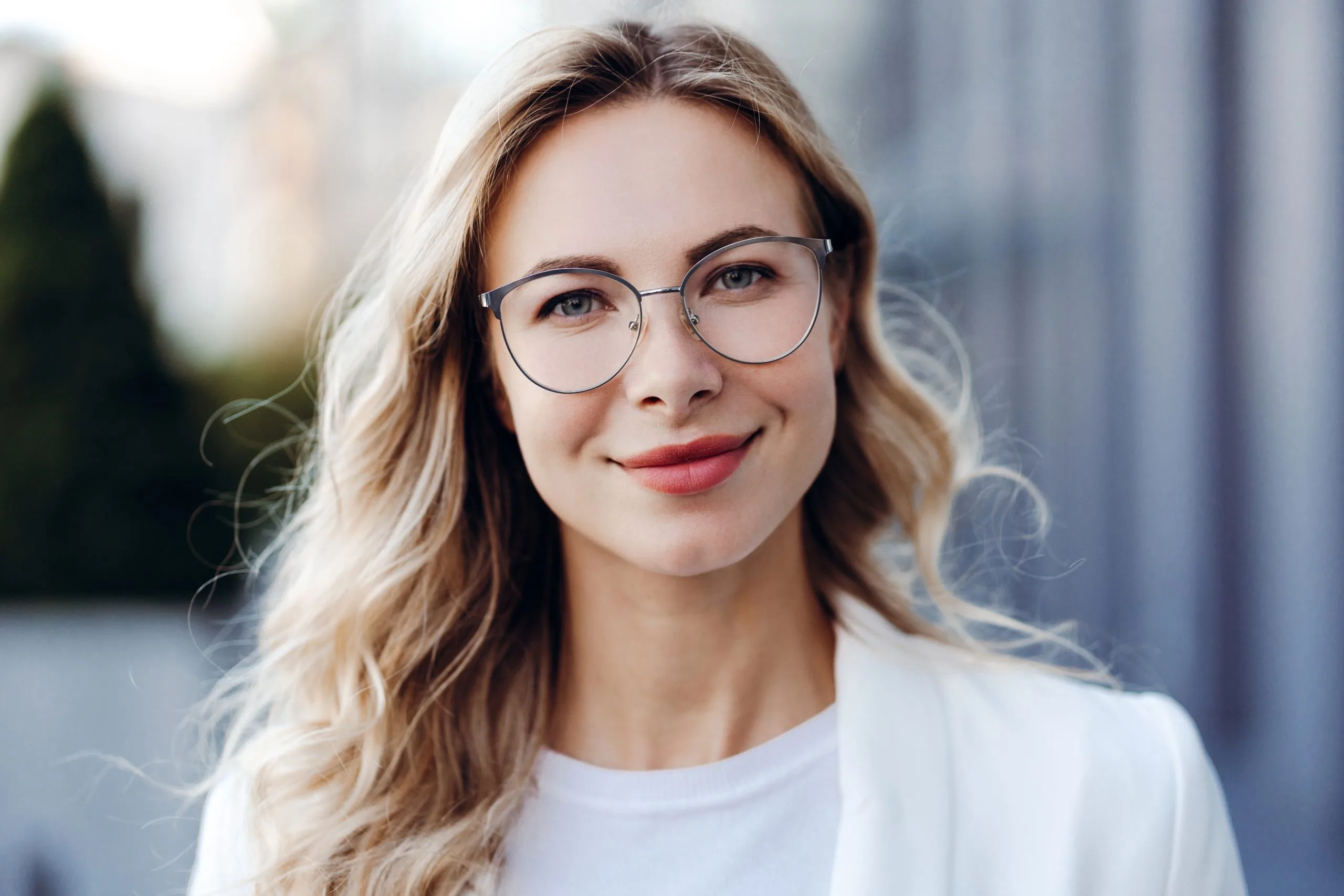 Woman wearing glasses and white shirt smiling outdoors with blurred building backdrop
