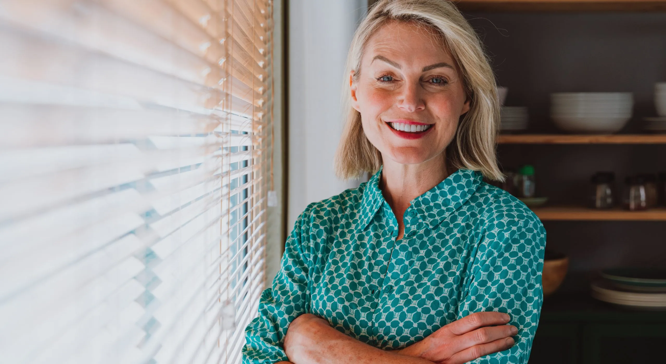 Woman with blonde hair smiling at camera in office with window blinds and shelving behind her