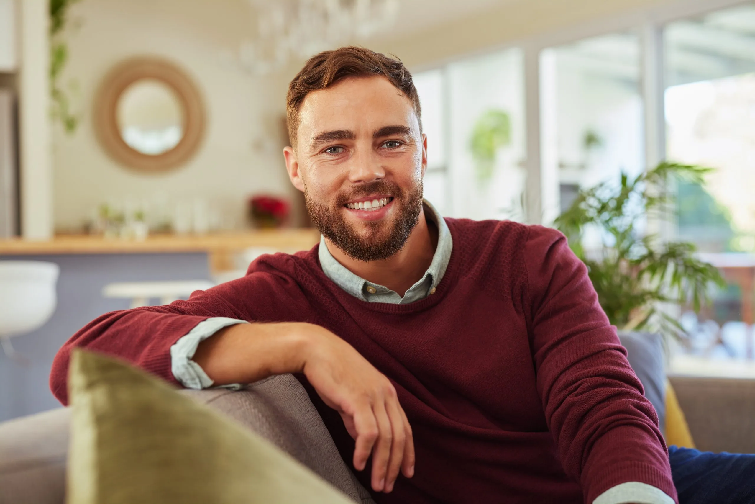 Smiling man in maroon sweater sitting on couch in modern living room with plants and natural light
