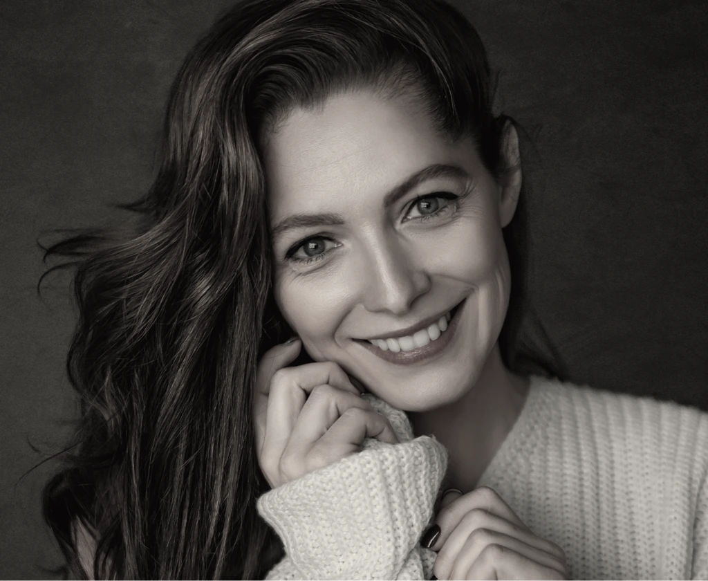 Woman with wavy dark hair smiling with hand near face wearing textured sweater