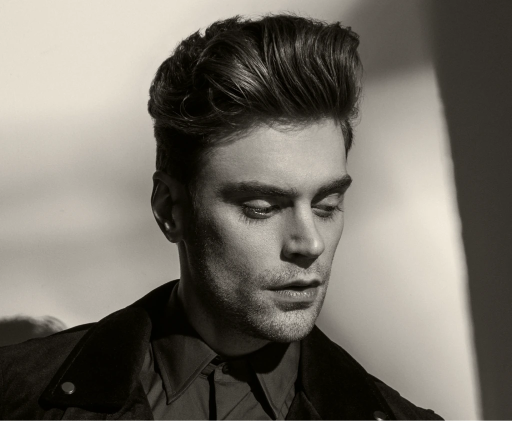 Man with styled dark hair in profile wearing dark shirt against neutral background