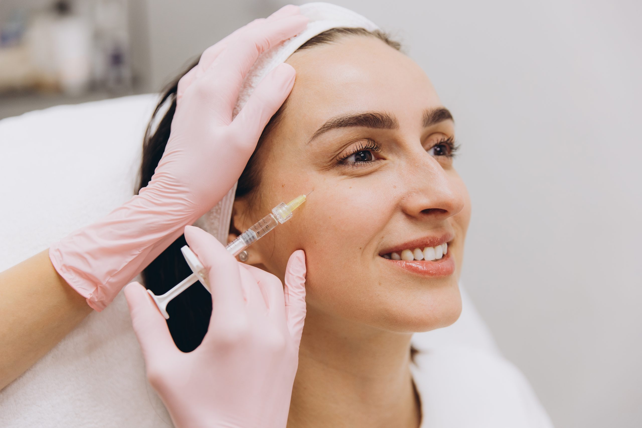 Aesthetician in pink gloves injecting dermal filler into patient's cheek during facial treatment