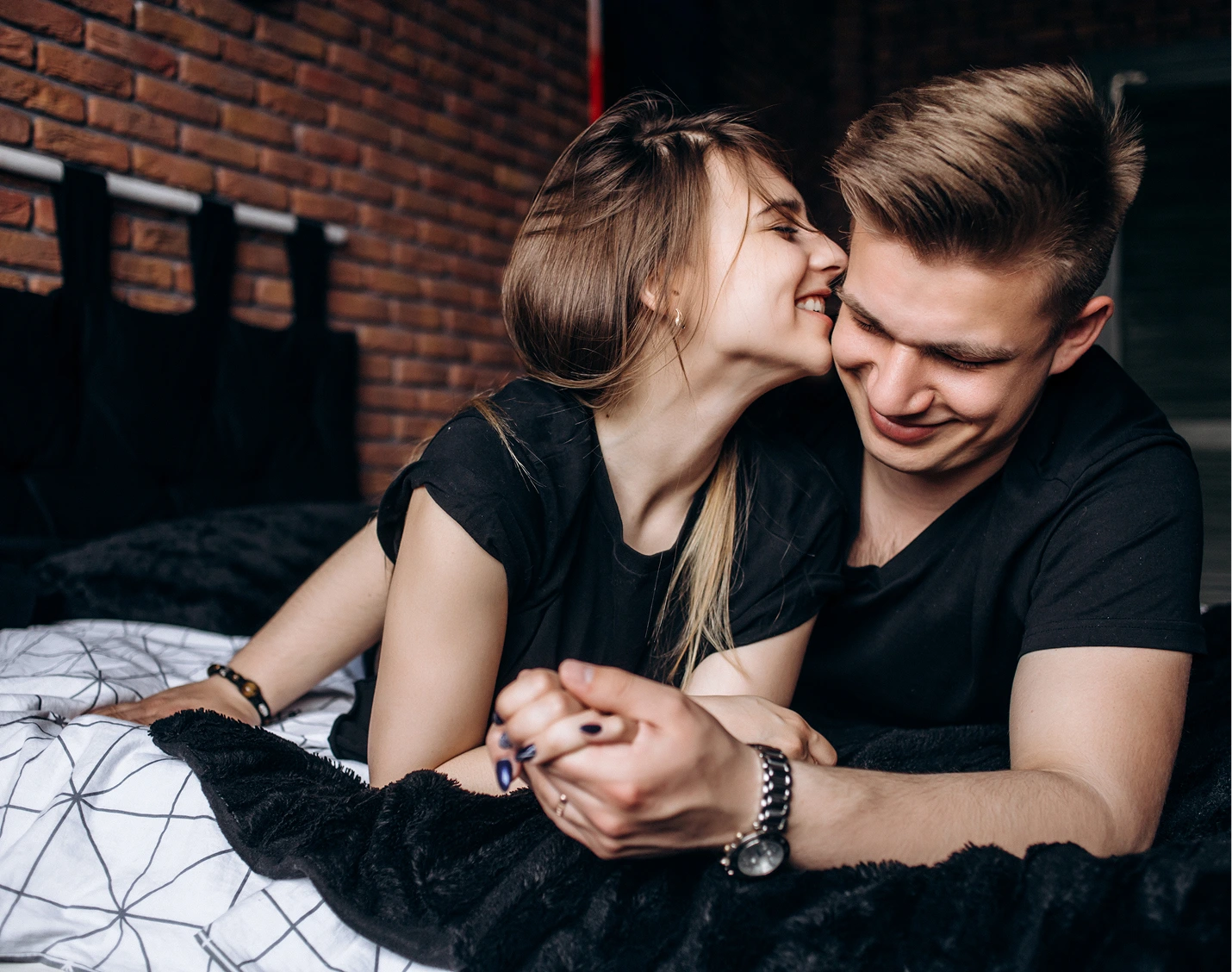 Couple in black shirts sitting on bed, woman kissing man's cheek while holding hands