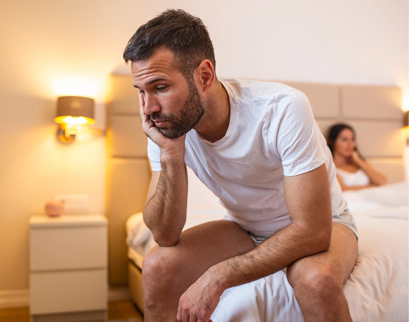 Man in white t-shirt sitting on bed with concerned expression, woman blurred in background