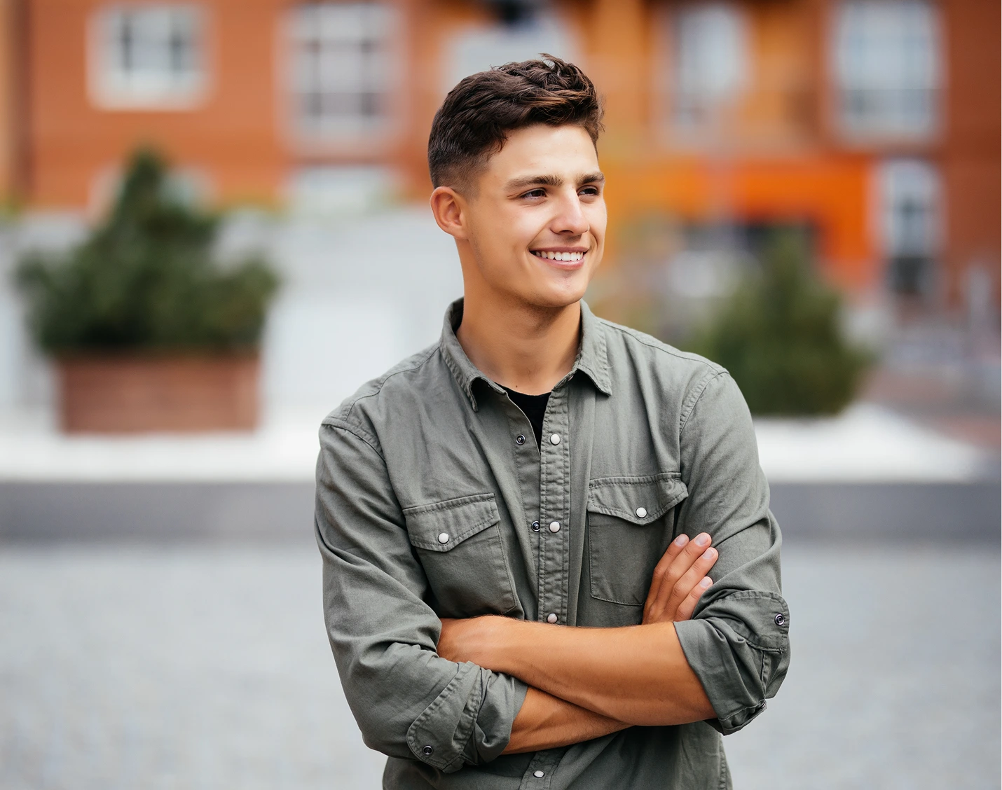 Young man in gray shirt smiling with arms crossed in urban courtyard