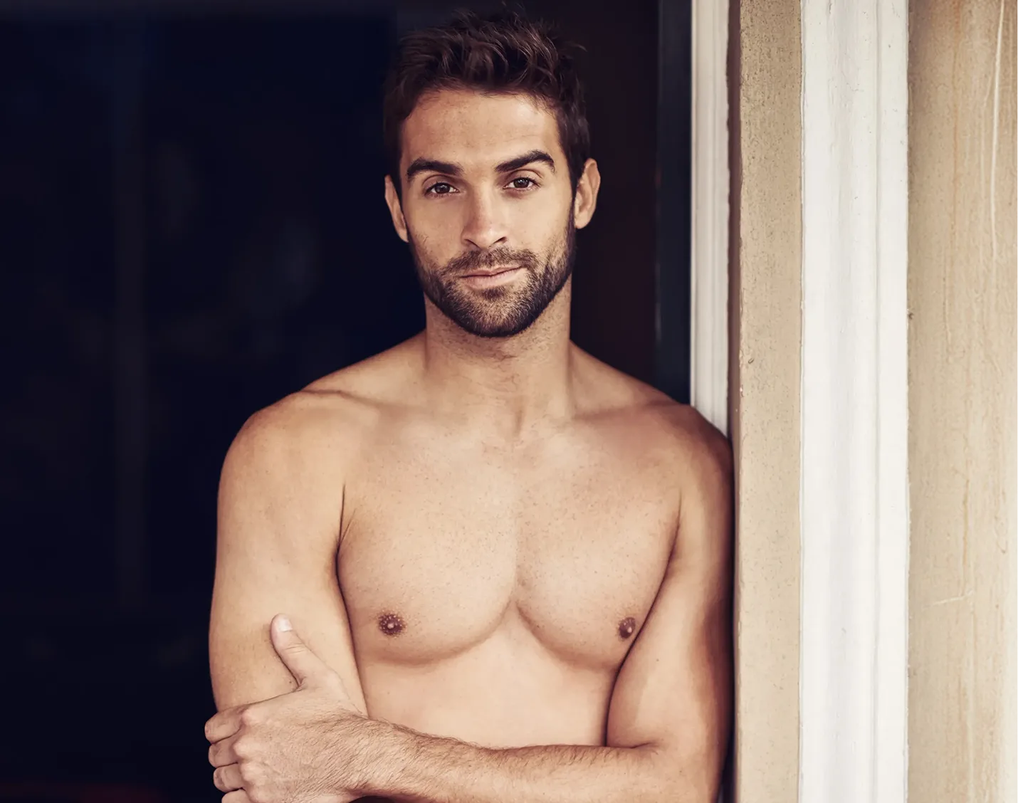 Shirtless man with sculpted chest facing camera in doorway with neutral background