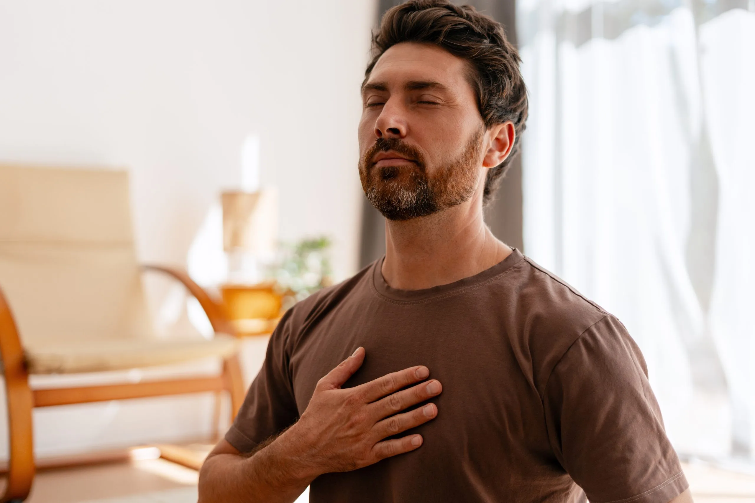 Bearded man in brown shirt with hand on chest, practicing mindfulness indoors