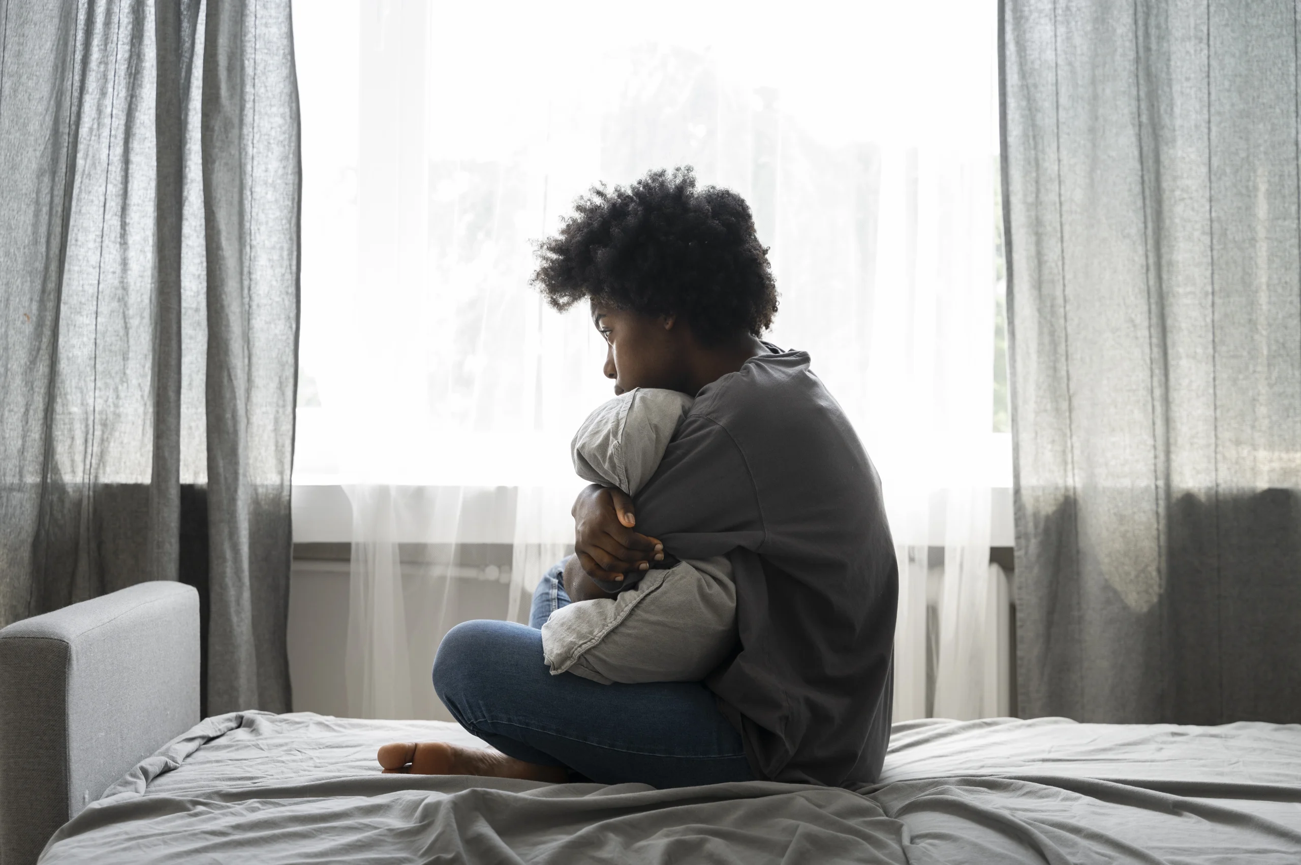 African American woman sitting awake on her bed hugging a pillow, looking out the window