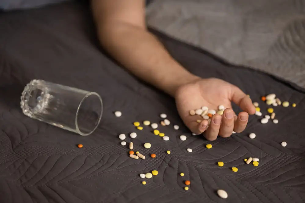 Person's hand holding scattered pills and capsules next to an overturned prescription bottle on dark fabric