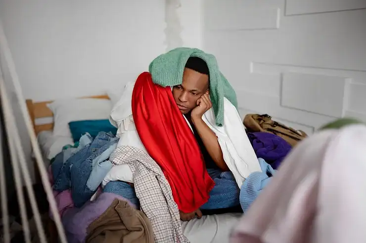 Person sitting on a bed surrounded by scattered laundry, wrapped in blankets, appearing withdrawn and distressed