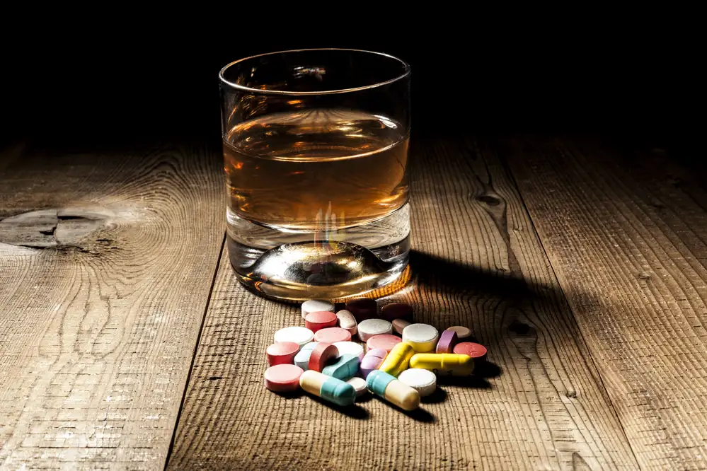 Glass of whiskey with scattered prescription pills and capsules on wooden surface