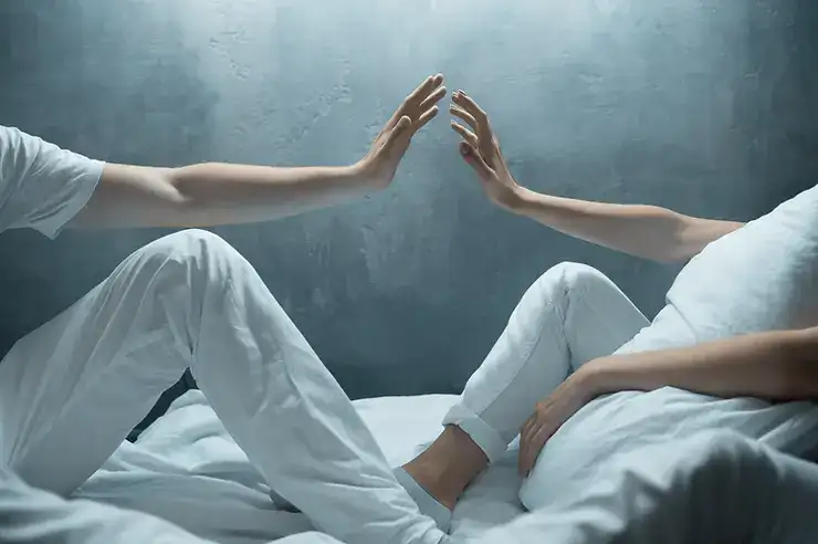 Two people in white clothing sitting on a bed with hands touching, facing each other
