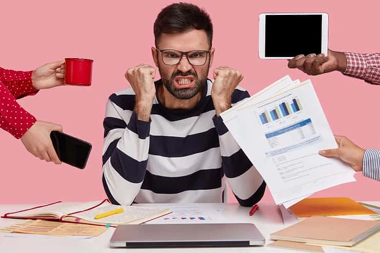Man in striped shirt showing stress with clenched fists while surrounded by work demands: coffee cup, phone, tablet, and charts