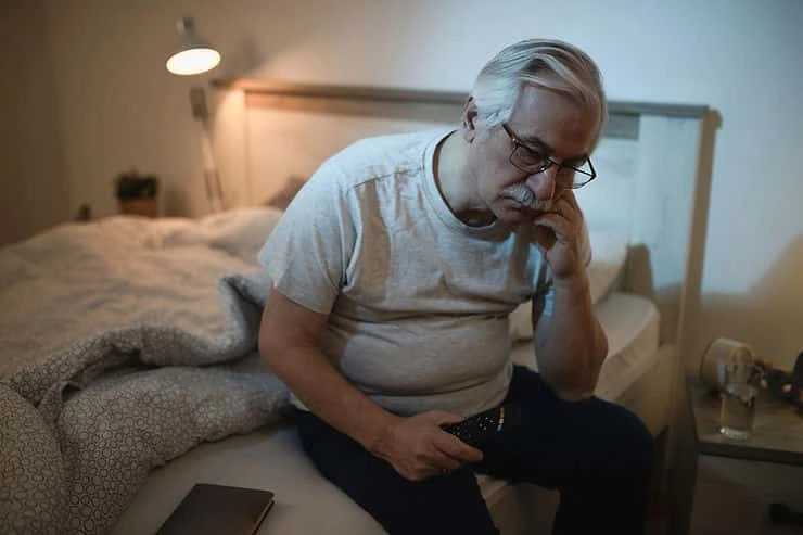 Older man sitting on bed at night, looking troubled and holding his head, depicting insomnia struggles
