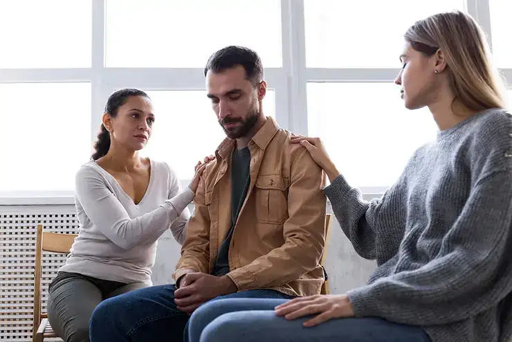 Two women and a man sit together in conversation near a bright window, depicting a supportive discussion about mental health challenges