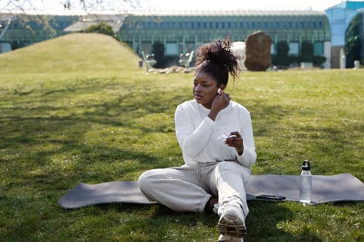 Woman in white clothing sitting on a yoga mat outdoors, reflecting peacefully in a green park setting