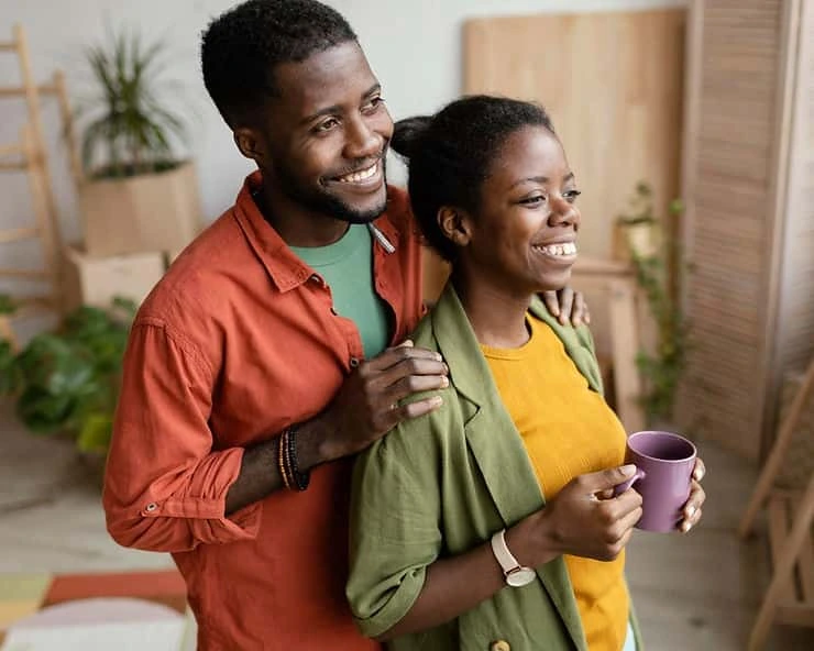 Smiling couple embracing in a bright home with potted plants, both holding mugs