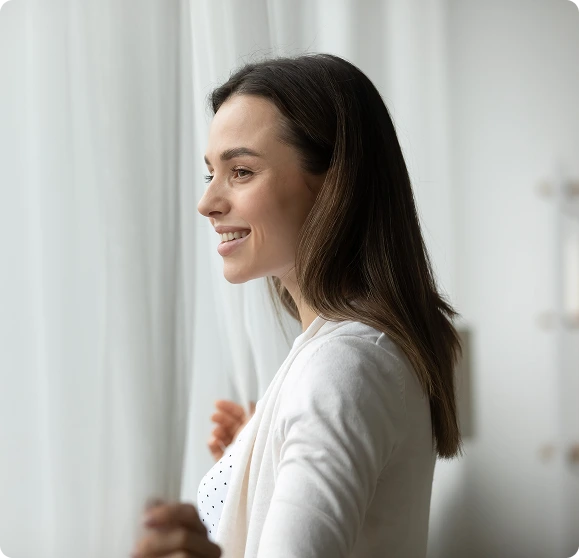 Woman smiling while looking out a window with sheer curtains