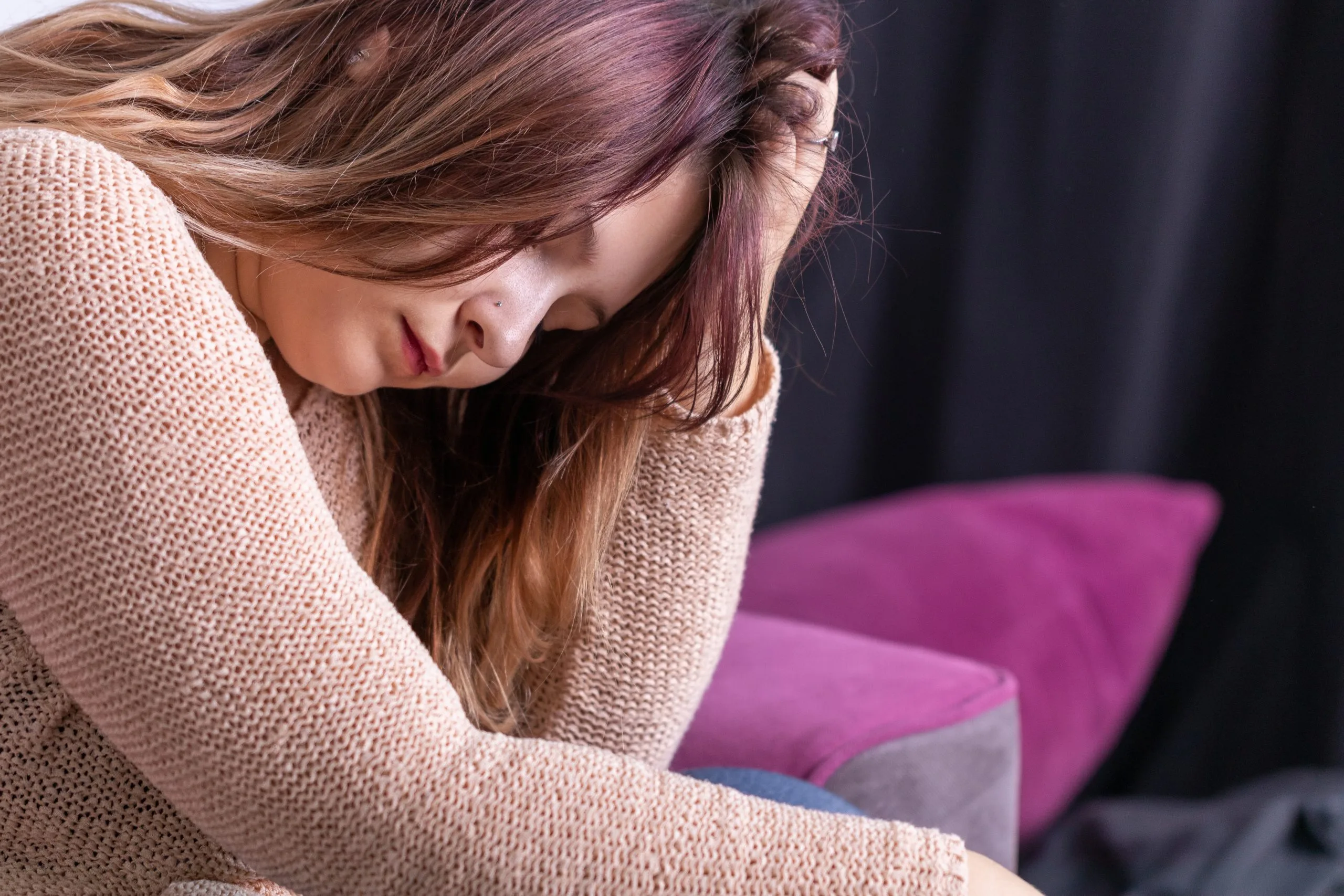 Woman with long brown hair looking down with a distressed expression, sitting on a purple chair