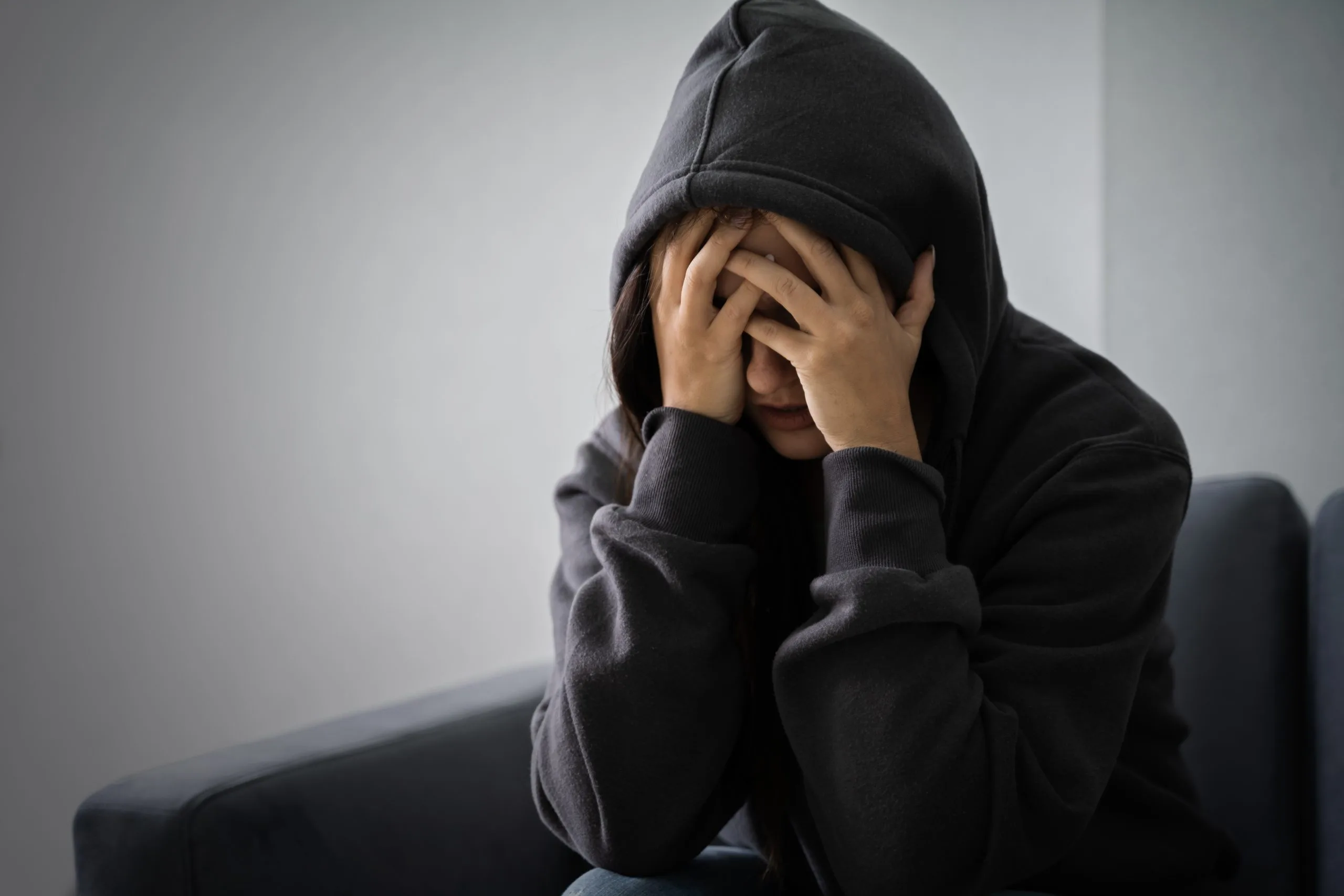 Woman in black hoodie sitting on sofa with head in hands, depicting emotional distress