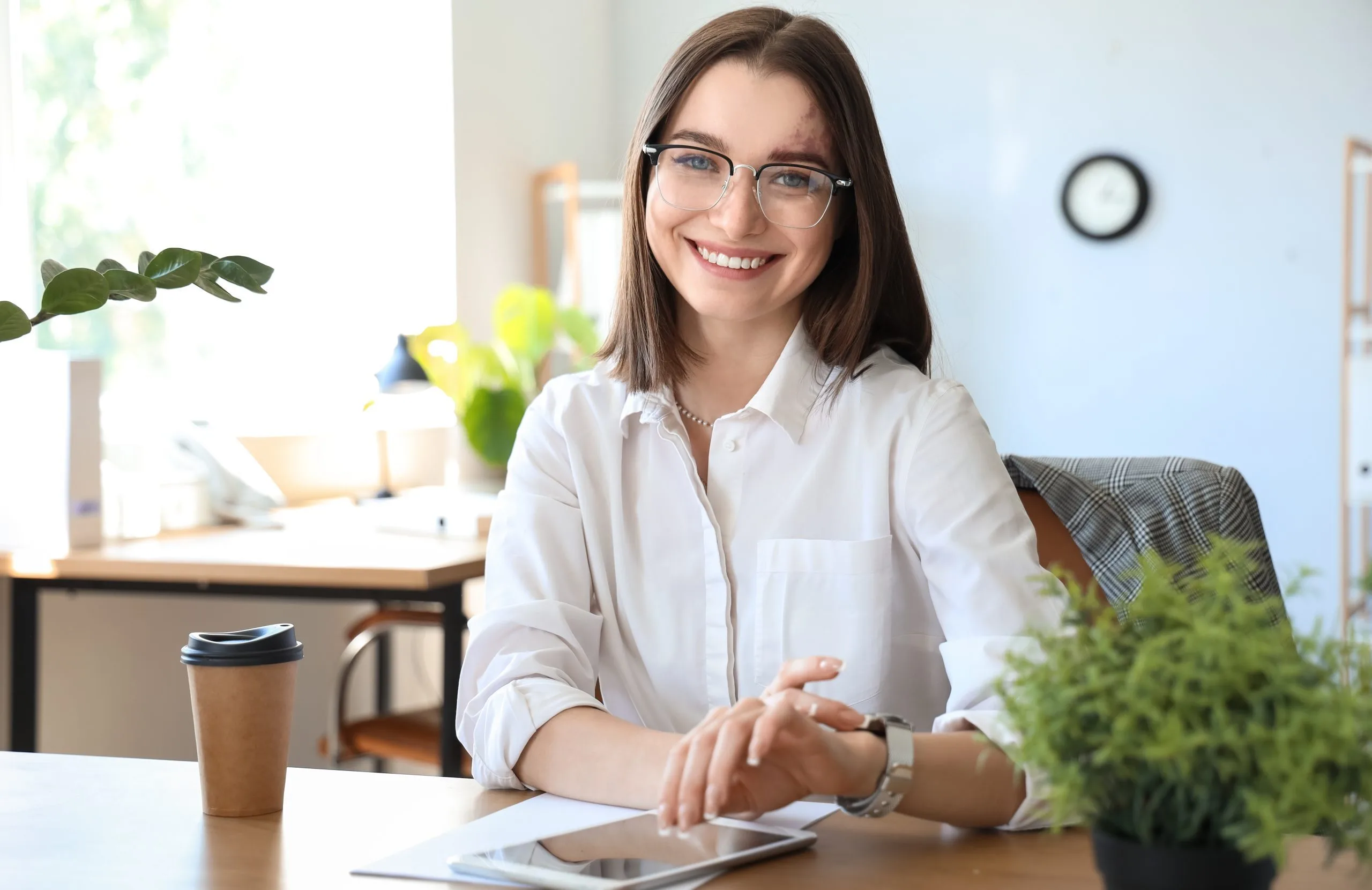 Woman in office setting smiling at desk with tablet and coffee cup
