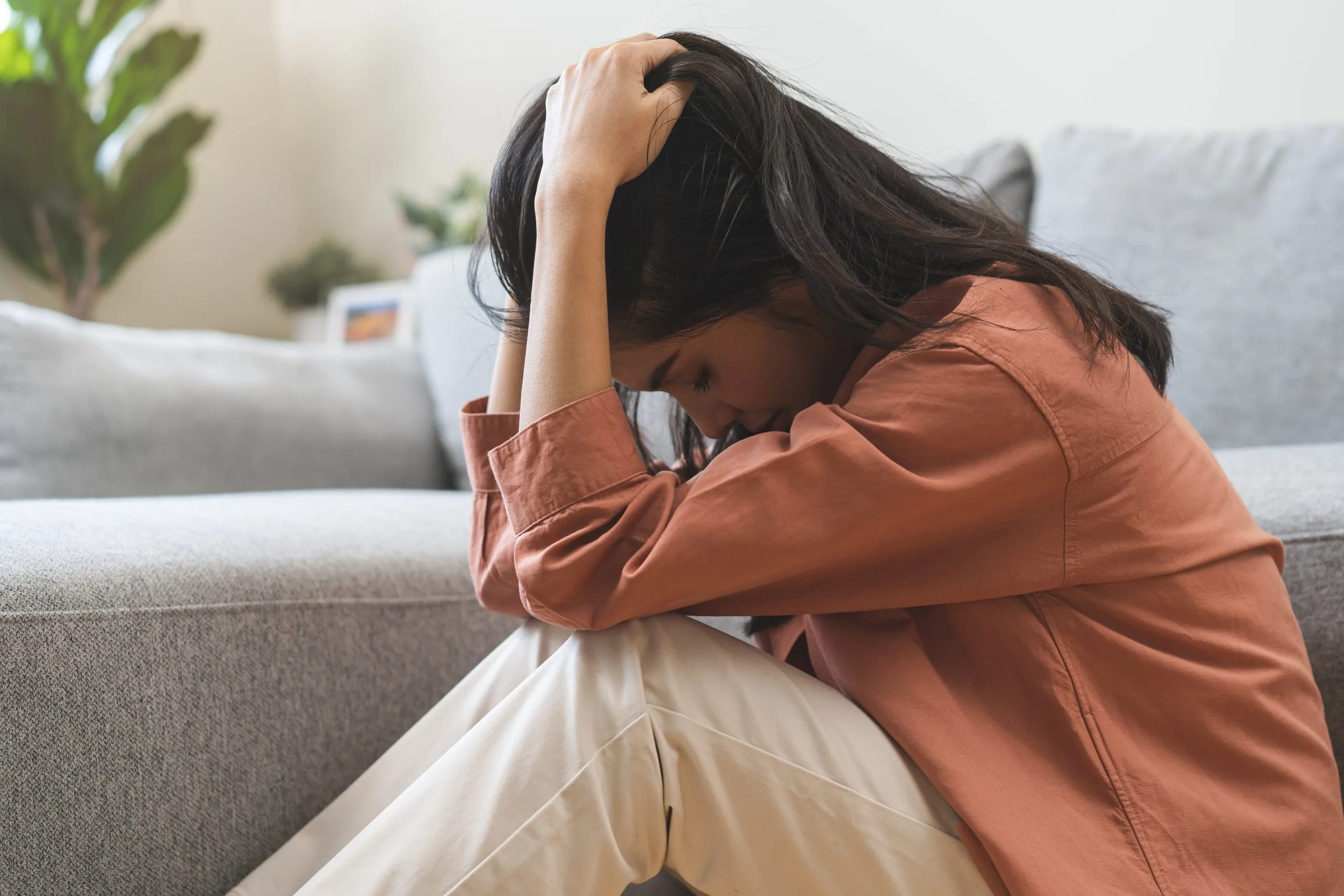Woman sitting on a couch with head bowed and hand covering face, expressing distress